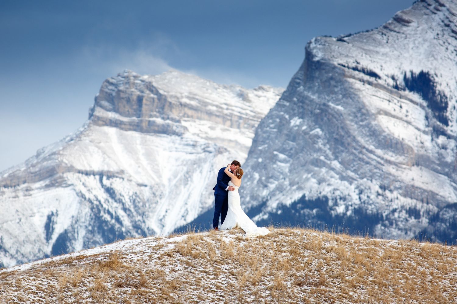 A couple shares a romantic moment on a mountain peak with snow-capped Banff mountains creating a majestic backdrop. Canmore three Sisters.