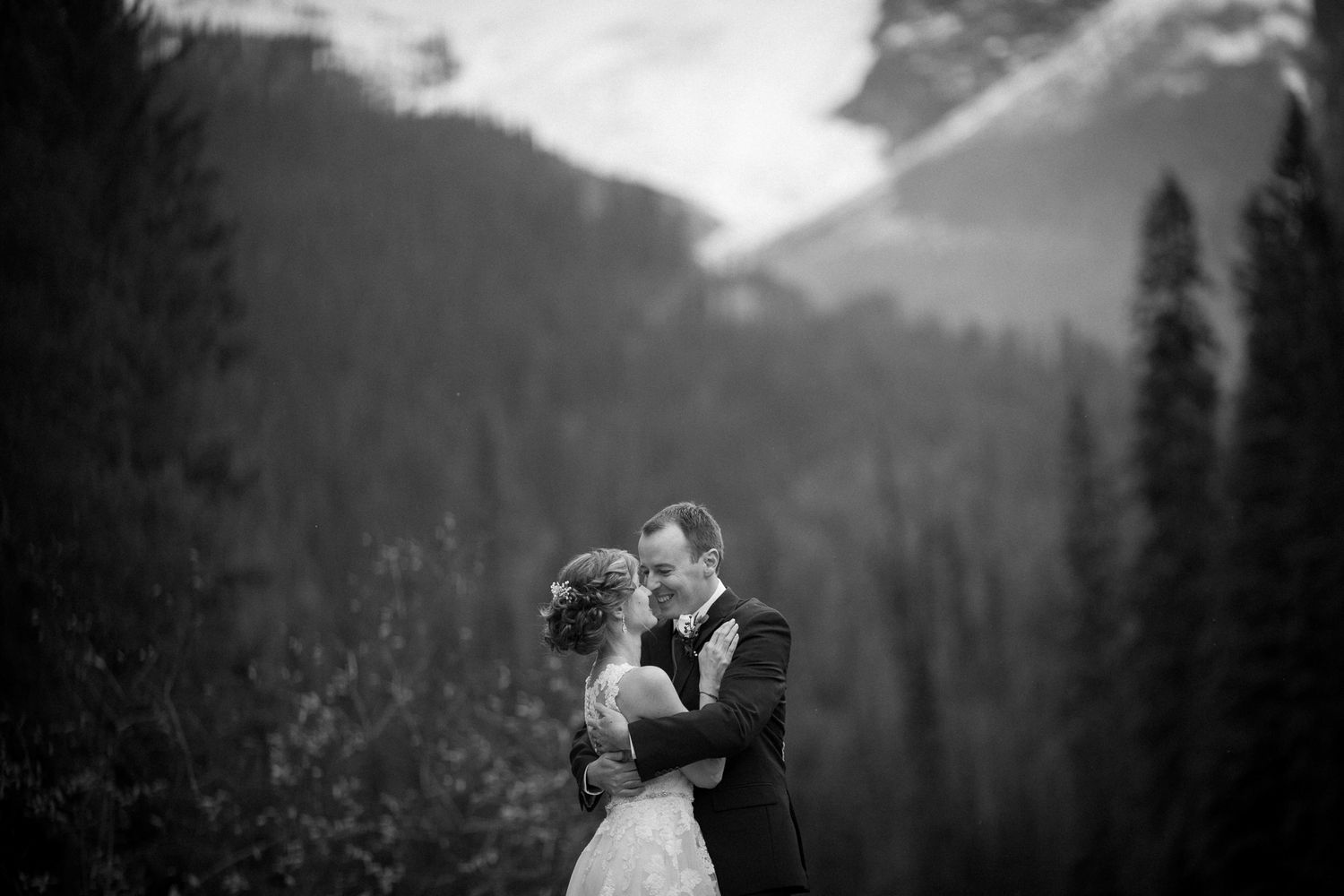 A couple shares a romantic kiss in front of a scenic mountain landscape during their wedding celebration. Emerald Lake Lodge, Field BC