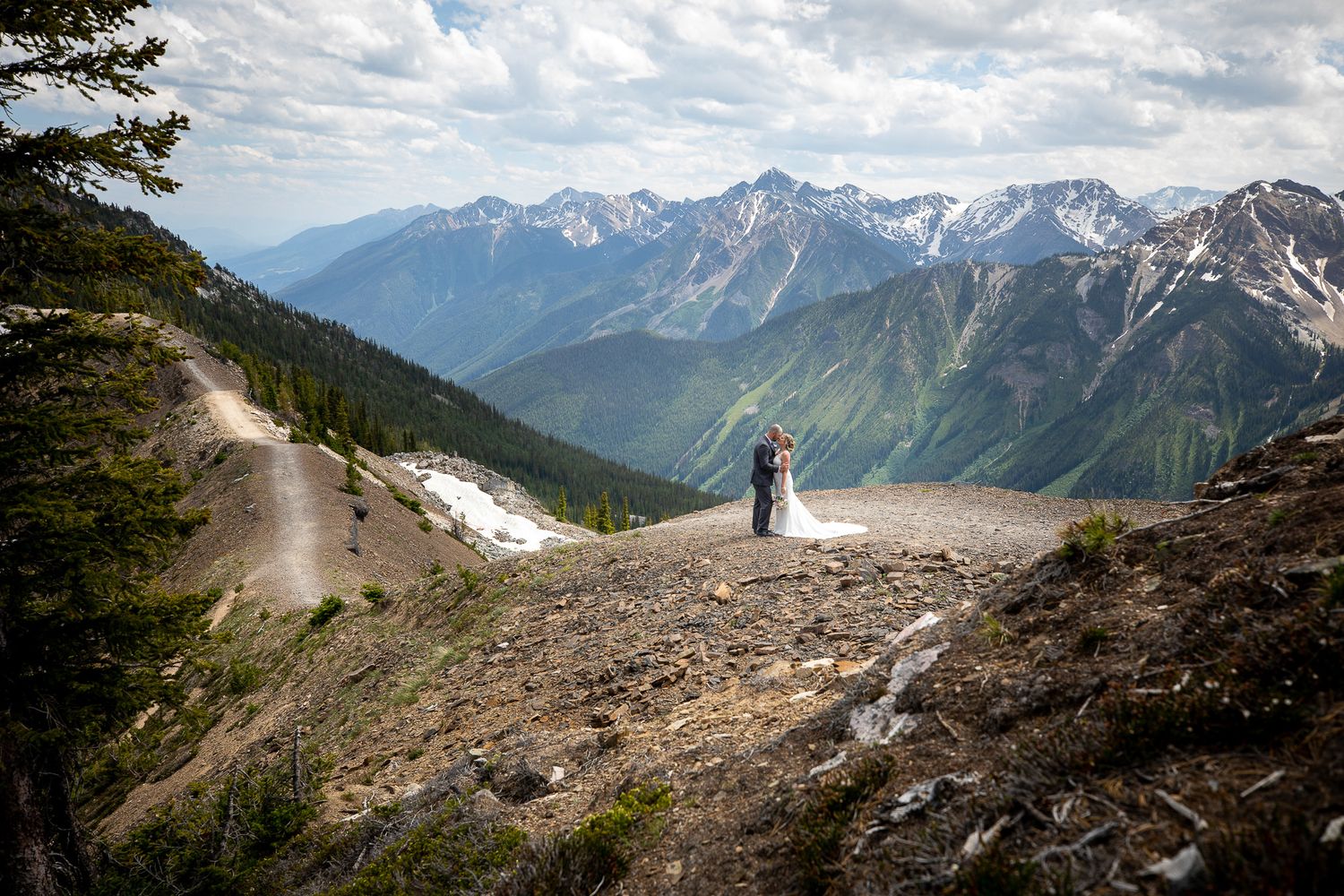 wedding couple on a scenic mountain trail winds through rugged alpine terrain with snow patches and distant peaks in Golden, BC.