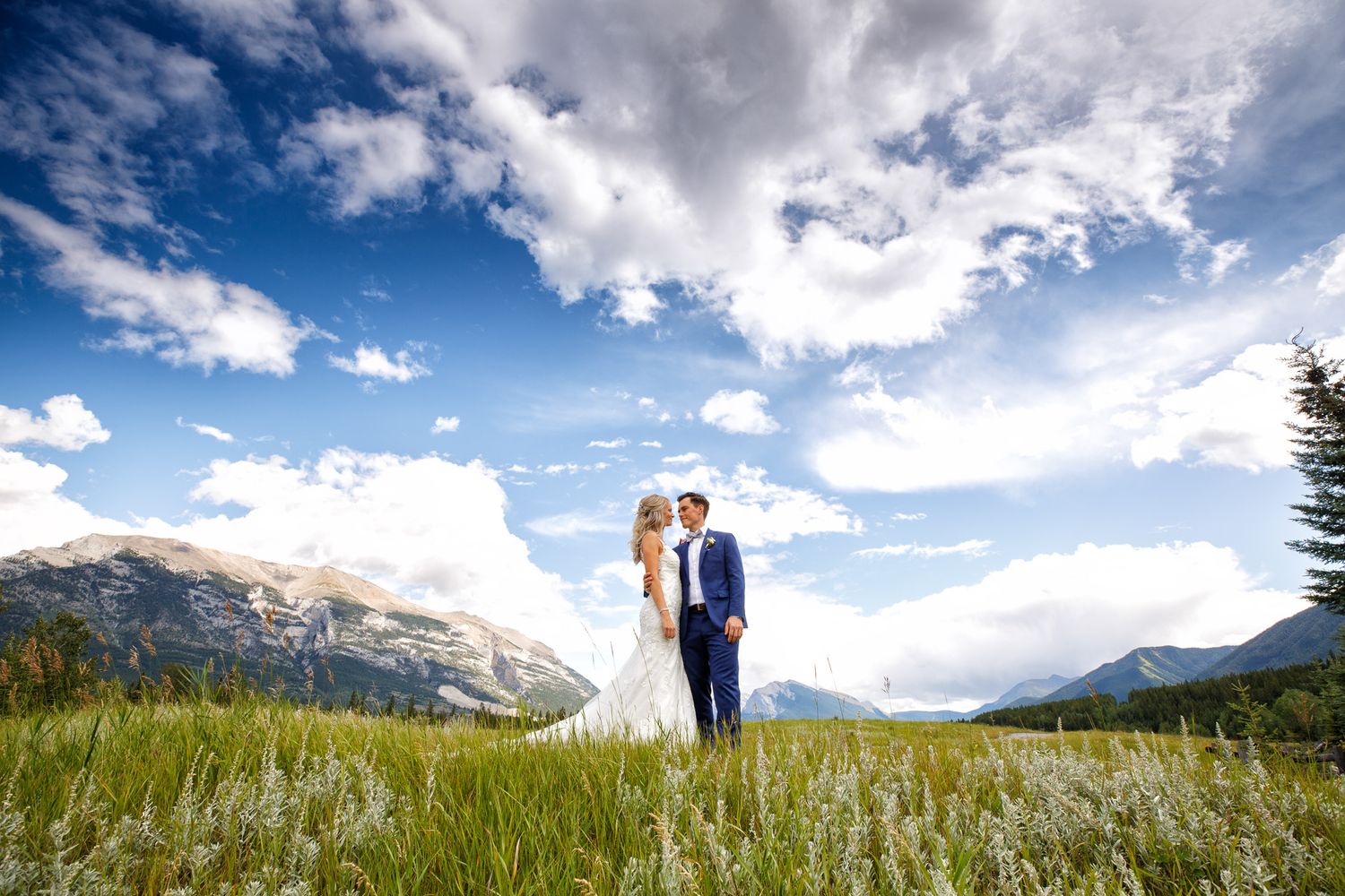 A couple stands together in a mountain meadow with dramatic clouds and peaks in the backdrop during a wedding photoshoot in Canmore.