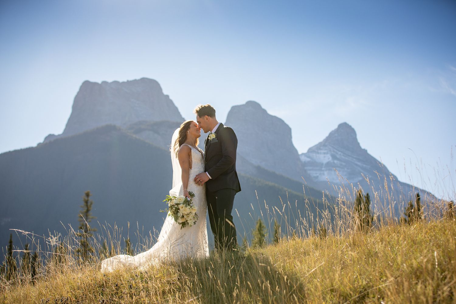 A couple shares a romantic moment in the mountains with dramatic peaks and golden grass in the foreground. Three Sisters Canmore.