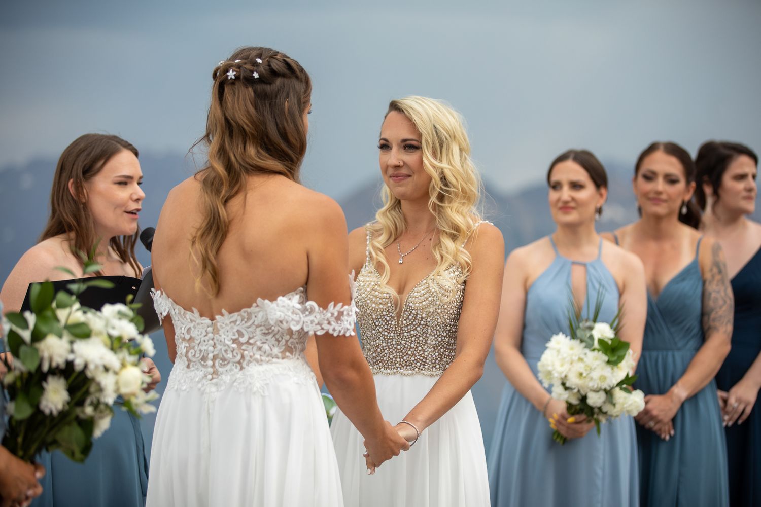 Two brides hold hands at outdoor wedding ceremony with bridesmaids in blue dresses holding white bouquets mat Kicking Horse Resort, BC