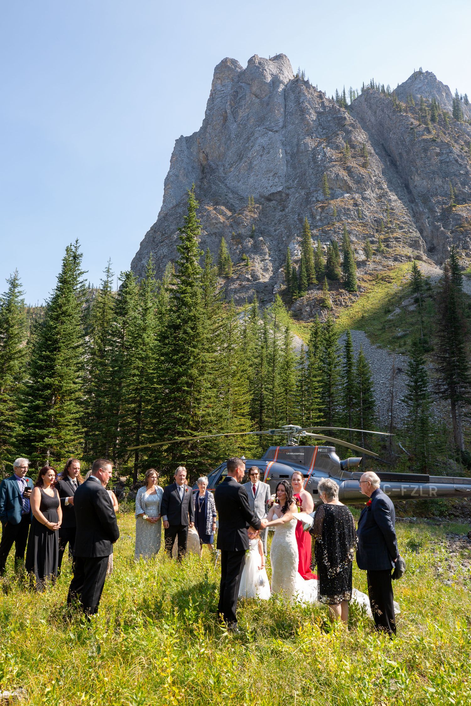 Small wedding ceremony in mountain meadow with helicopter and rocky peaks backdrop in Kananaskis, Alberta.