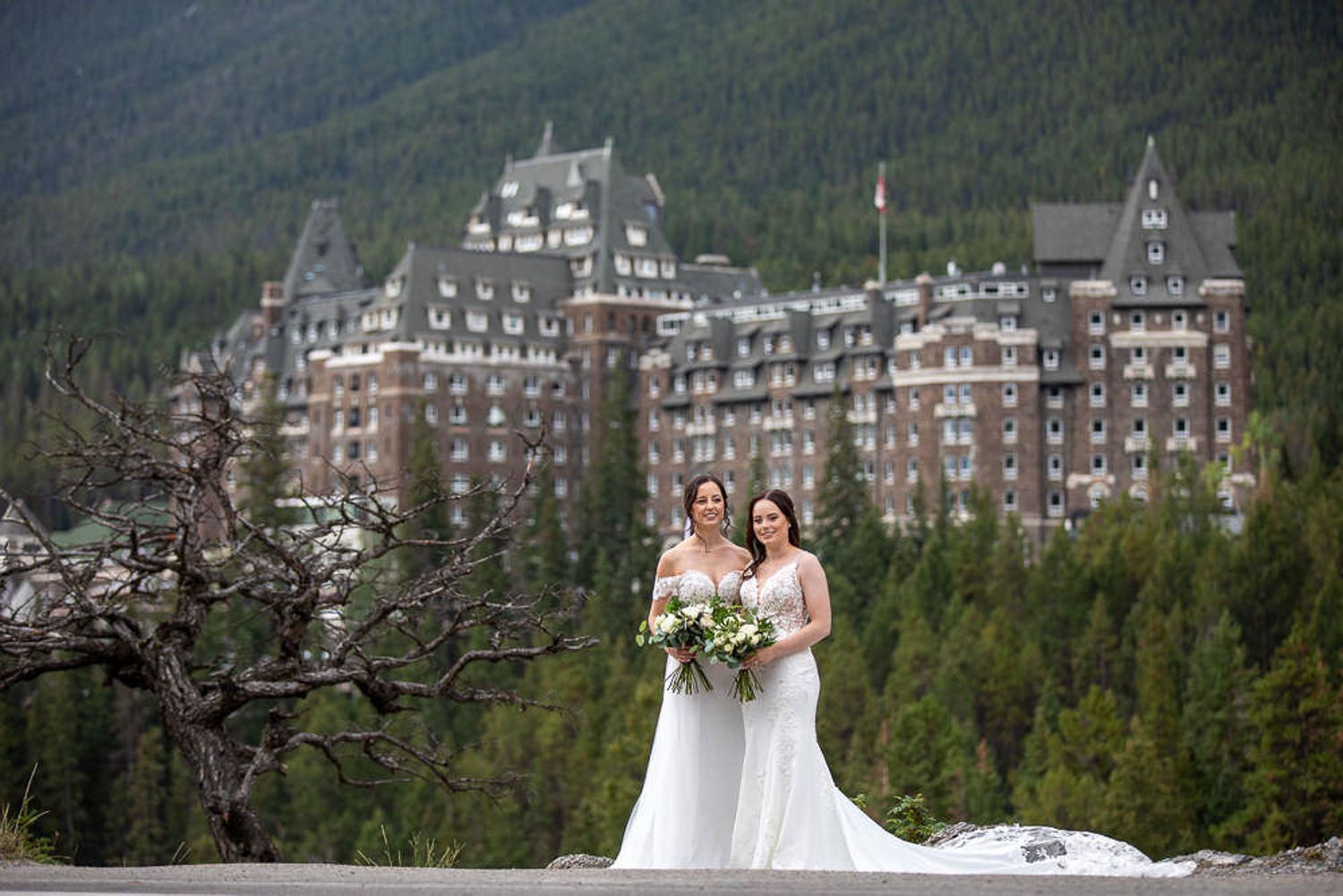 Two LGBTQ brides posing at surprise corner in Banff with the majestic Fairmont Banff Springs Hotel in the background.