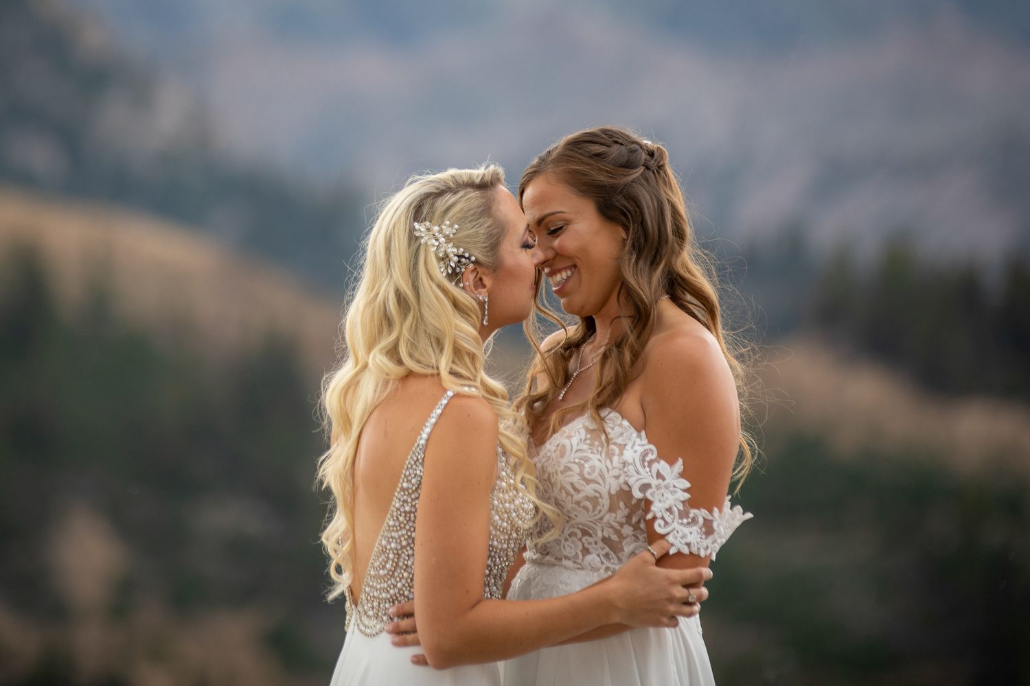 Two brides in wedding attire embrace against a scenic mountain backdrop during a sunset ceremony at Kicking Horse resort in Golden, BC