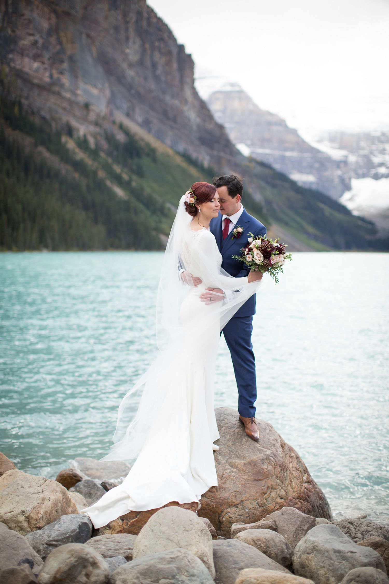 A romantic wedding couple stands on rocky shores of Lake Louise with turquoise waters and mountain peaks in the background.