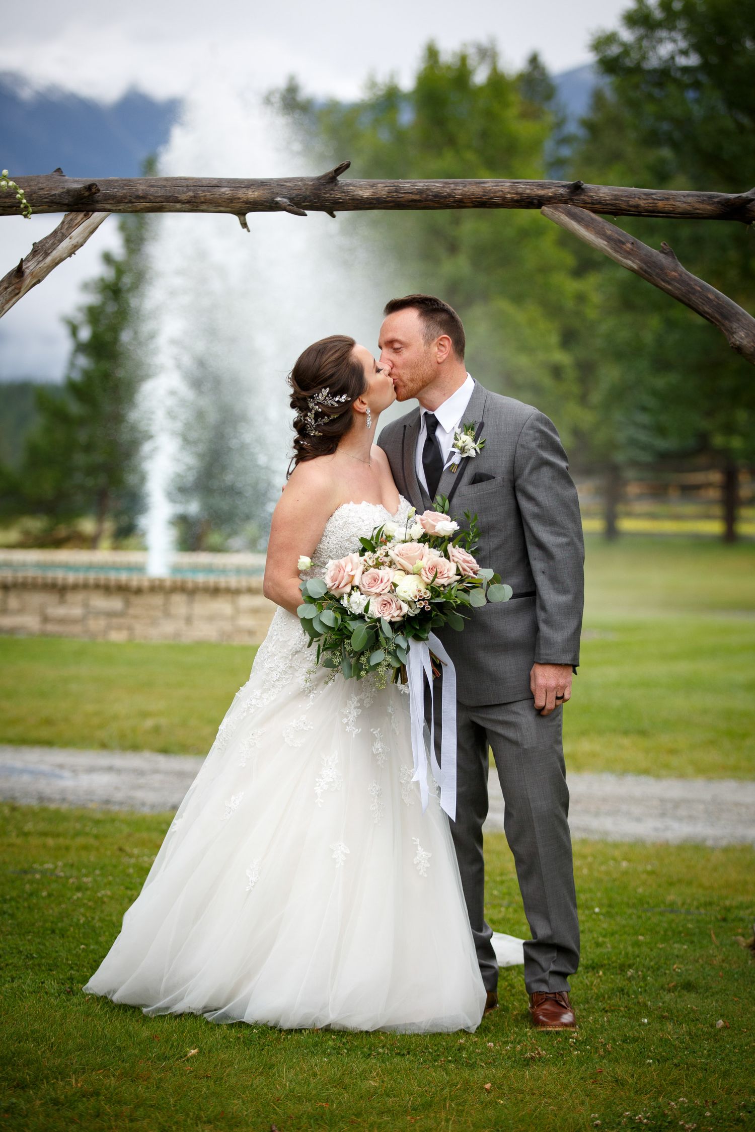 Bride and groom kissing under rustic wooden arch on green lawn with fountain and mountains in background at Cherry creek Estate in Kimberley