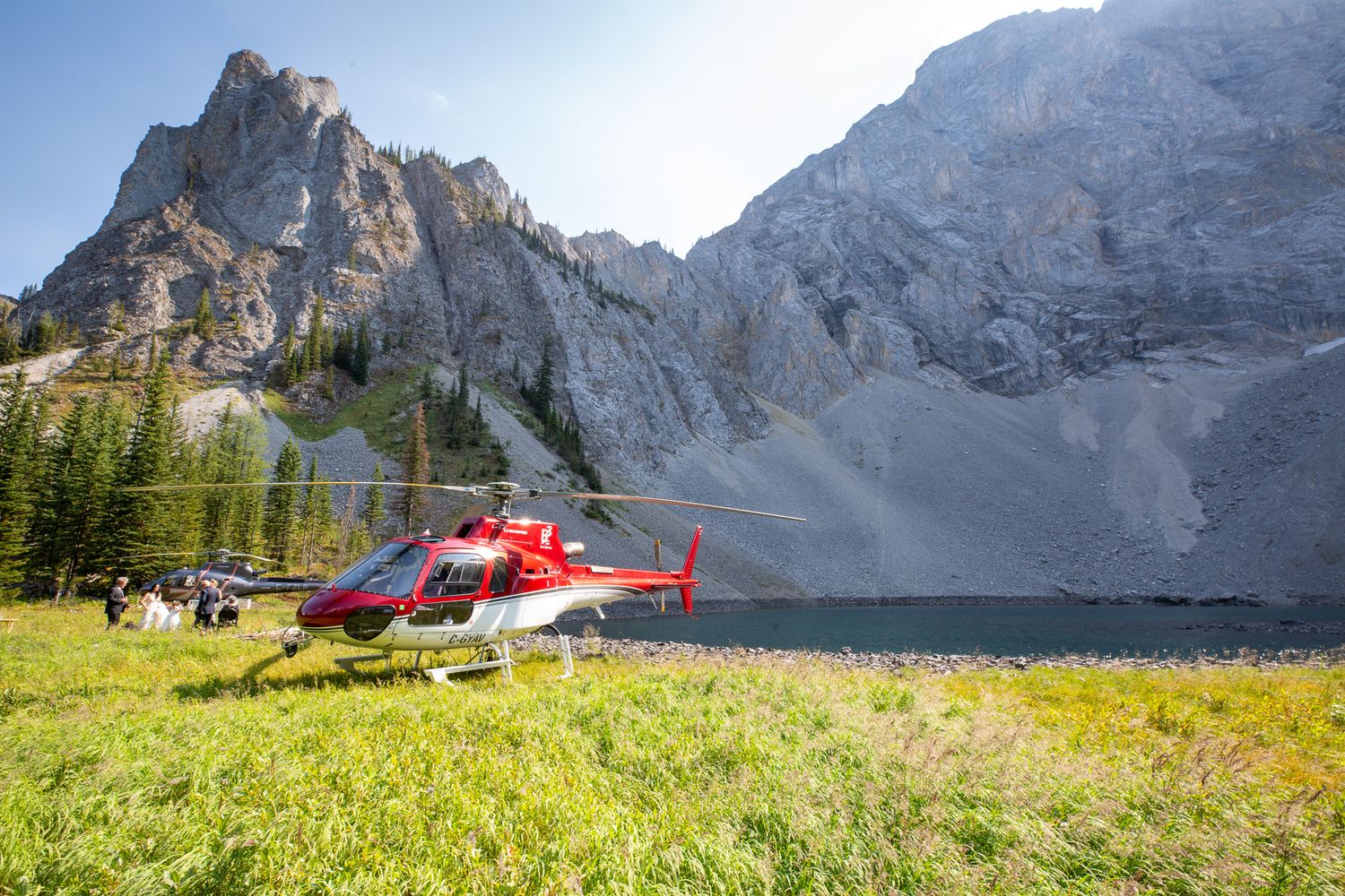 Red helicopter landed on alpine meadow beside mountain lake with rocky peaks and evergreen trees in background in Kananaskis, Alberta.