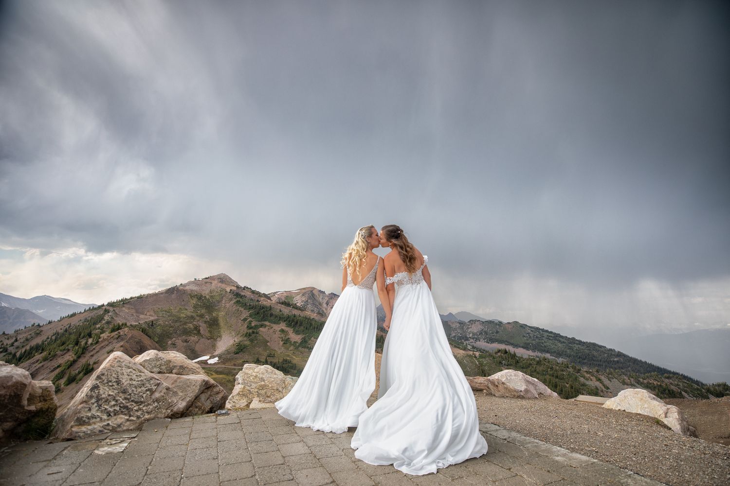 Two brides in white gowns embrace on a stone terrace with dramatic mountain and stormy sky backdrop at Kicking Horse Resort, Golden, BC