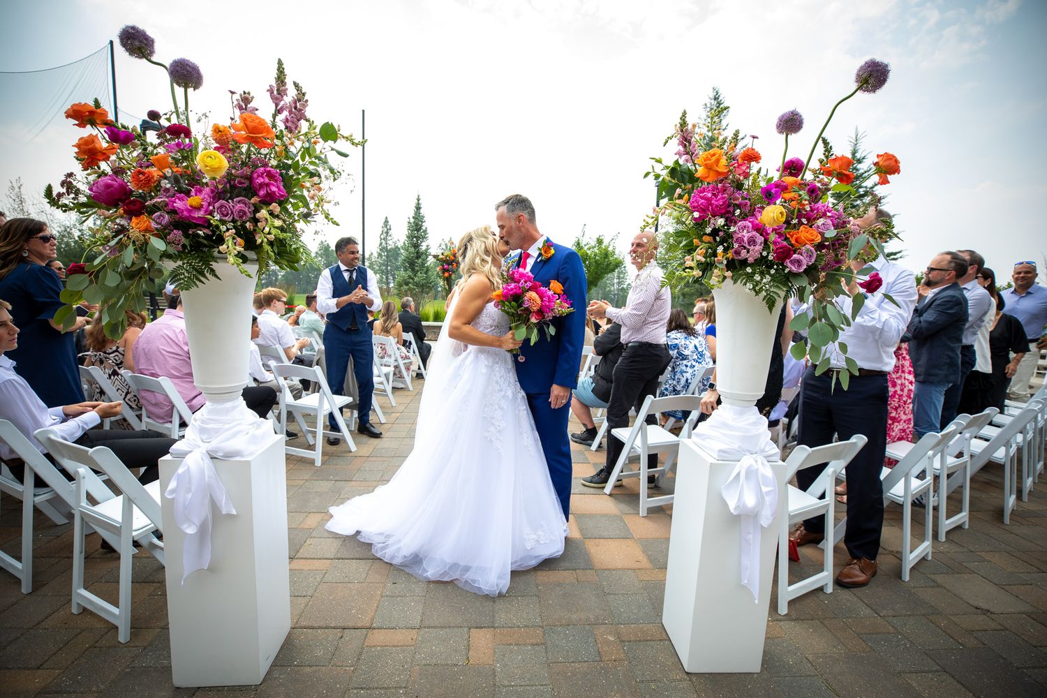 Bride and groom share a kiss at outdoor wedding ceremony with vibrant floral arrangements and seated guests. Copperpoint resort,  Invermere.