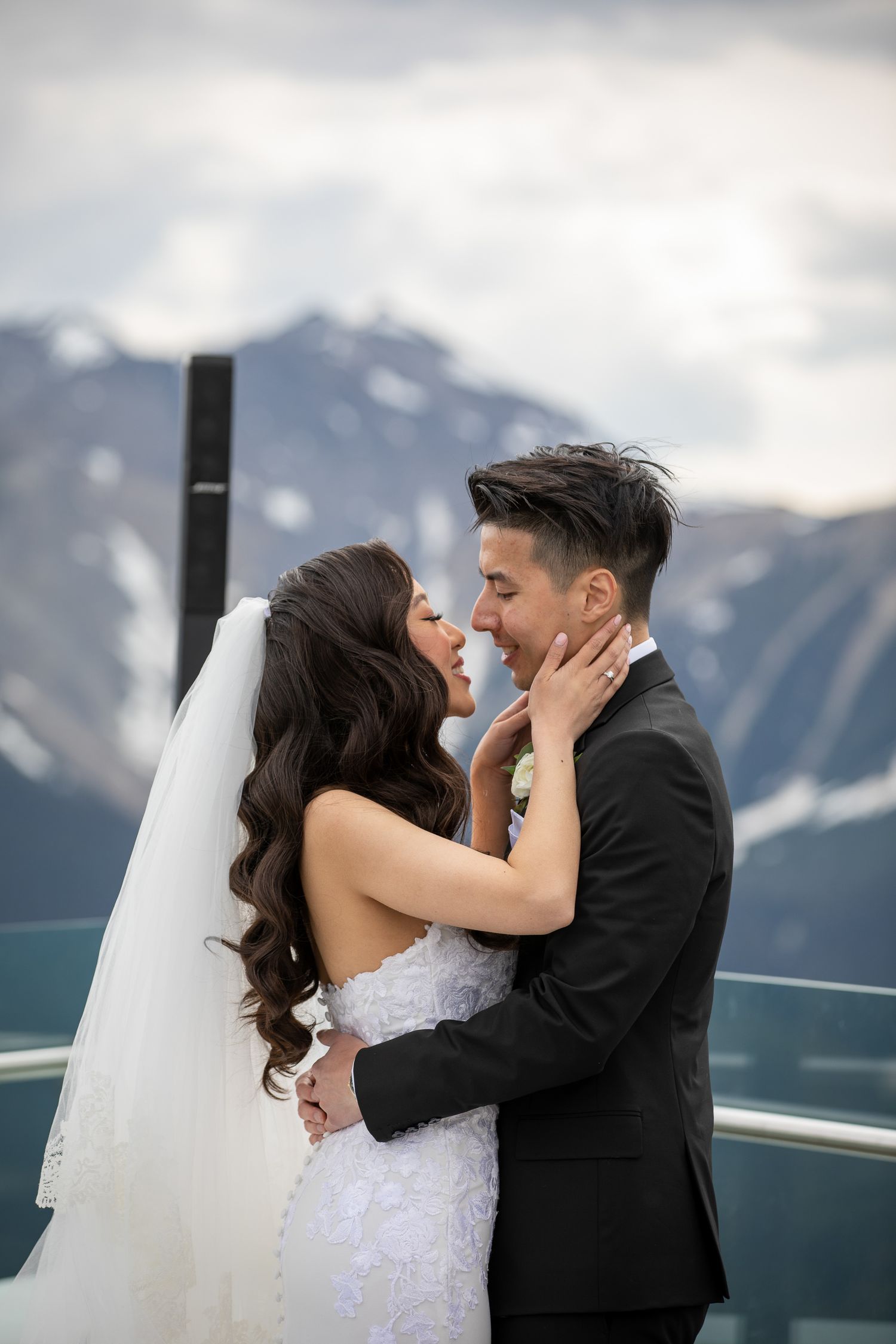 Bride and groom share a romantic moment on a mountain overlook, with snow-capped peaks in the background.