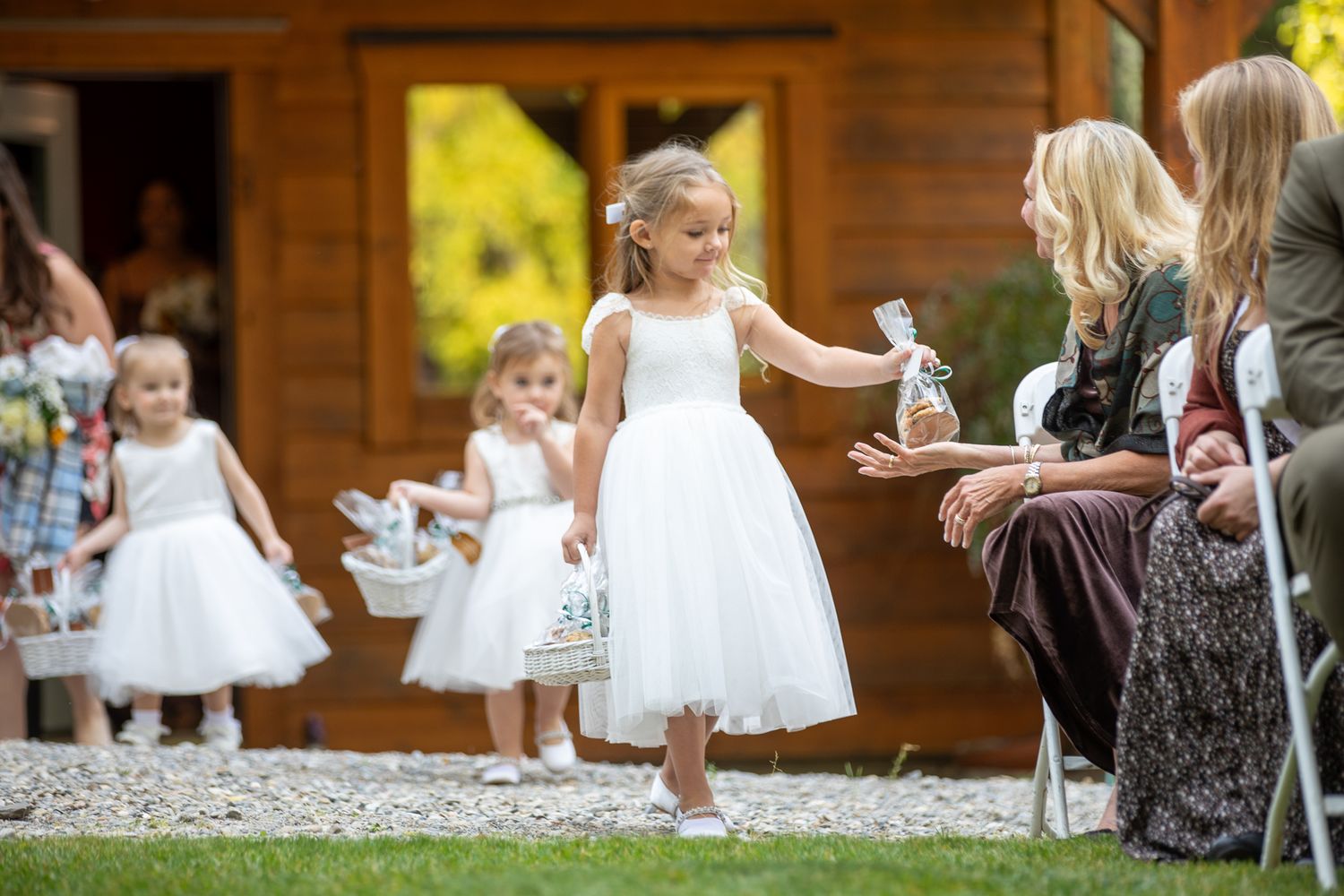 Young flower girls in white dresses dance and twirl at an outdoor wedding celebration with autumn colors in background.