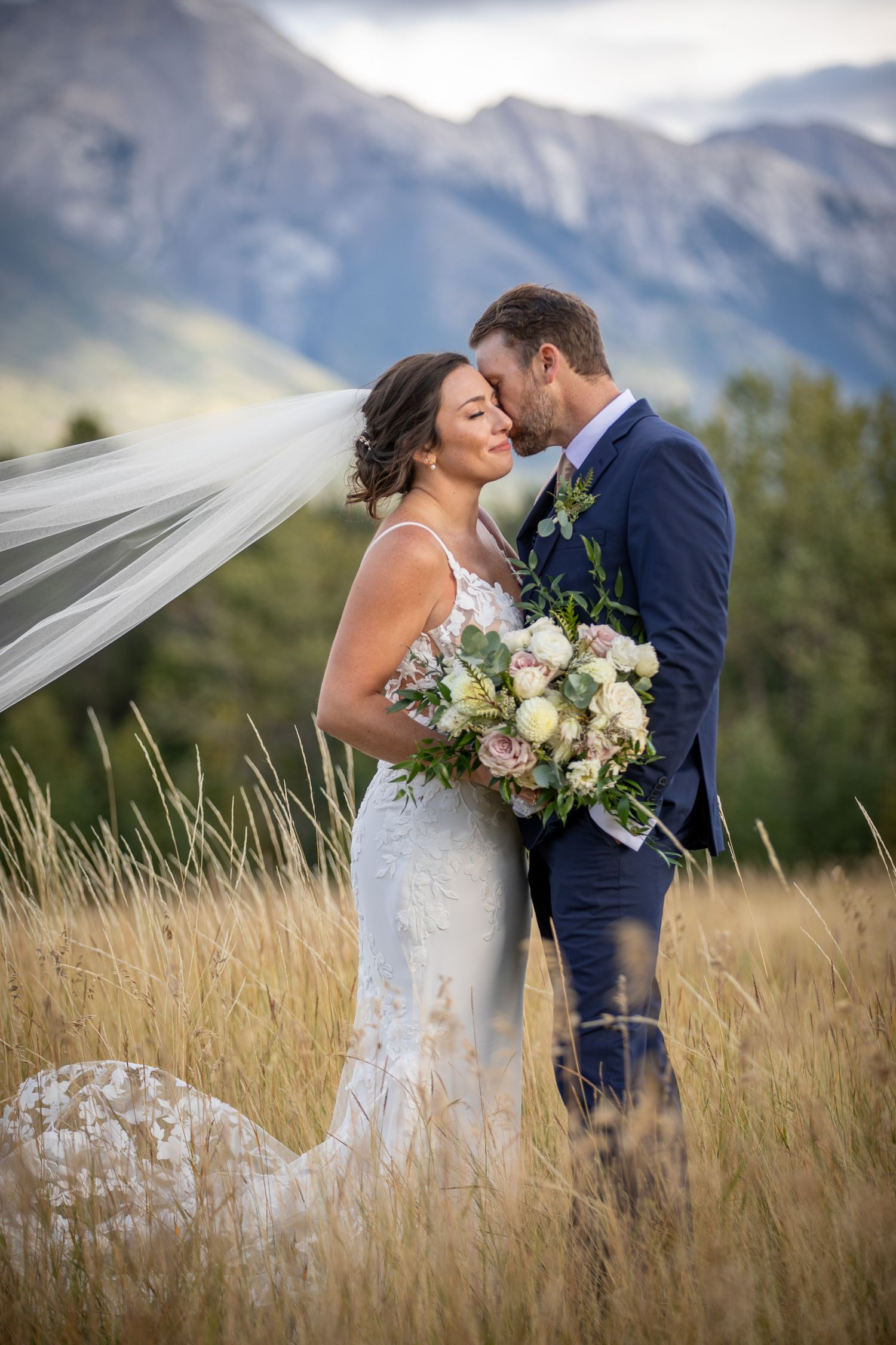 A romantic wedding couple embraces in a mountain meadow with flowing veil and white floral bouquet against mountain backdrop. Banff, Alberta
