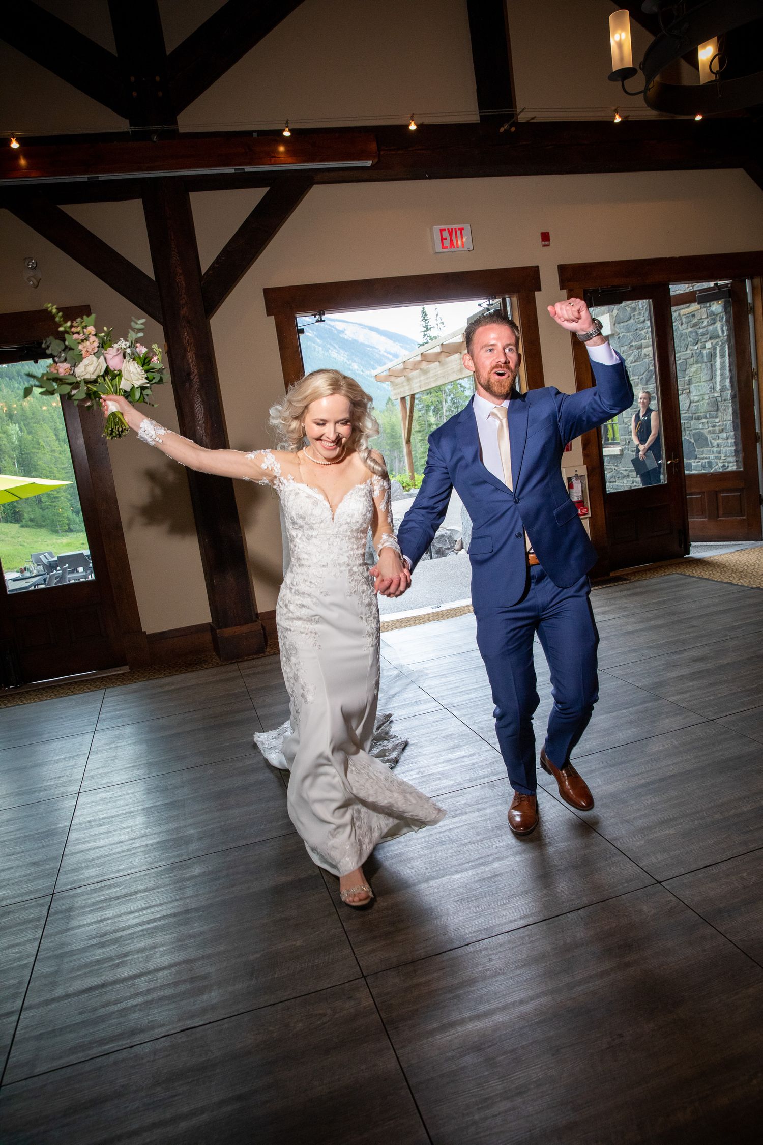 Joyful newlyweds make grand entrance at wedding reception, bride in lace gown holding bouquet, groom in navy suit cheering at Silvertip.