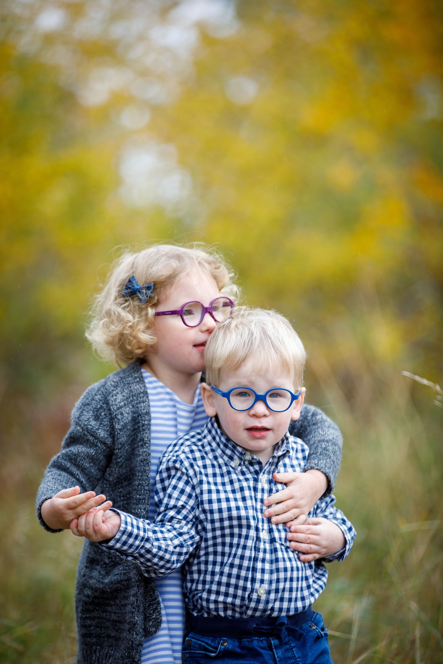 Two children wearing matching blue glasses and plaid shirts embrace outdoors against a blurred autumn background at Calgary Zoo.