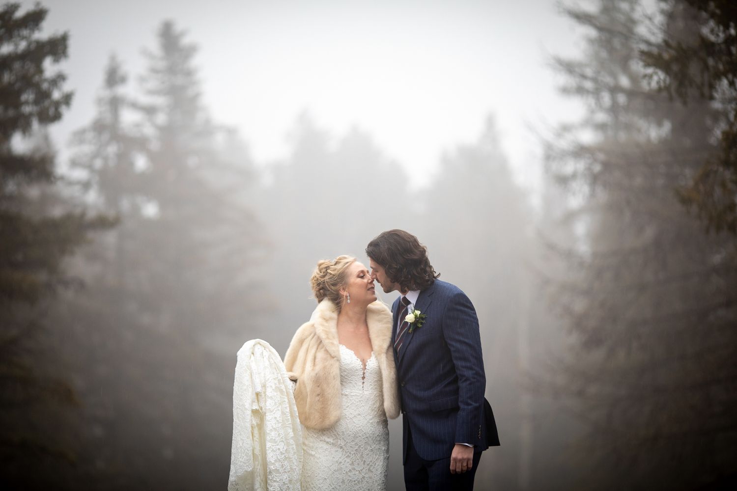 A couple shares an intimate moment in a misty forest setting wearing formal wedding attire at the Fairmont Banff Springs Hotel in Banff.