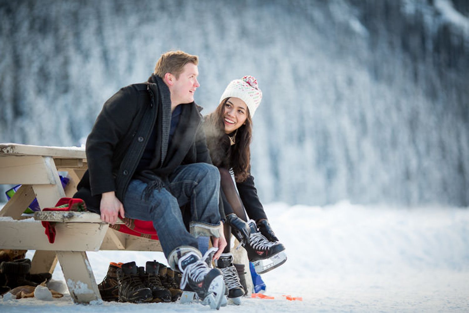 A couple sits together on a wooden bench in a snowy winter landscape with snow-covered mountains in the background in Lake Louise, AB.