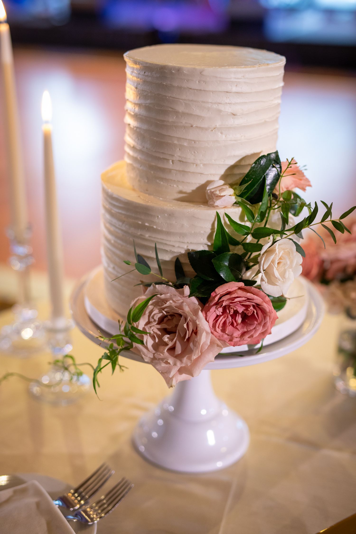 White textured wedding cake adorned with pink roses and greenery displayed on a white pedestal cake stand.
