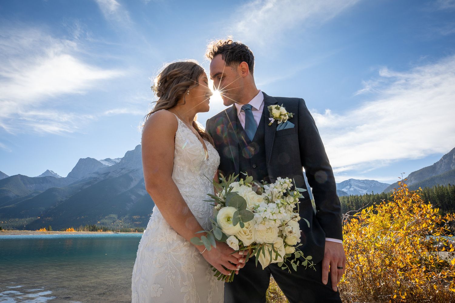 A bride and groom embrace in a scenic outdoor setting with mountains and a lake in the background at Quarry lake in Canmore, AB.