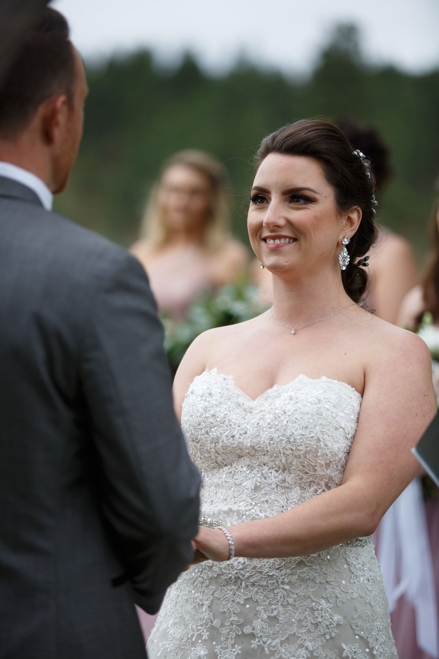 Bride in strapless lace wedding gown smiling at groom during outdoor wedding ceremony with bridesmaids in pink behind her.
