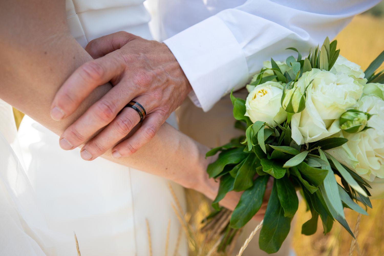 Newlywed couple holding hands with a black wedding ring and a white rose bouquet with green leaves at Swansea Ranch in Invermere, BC
