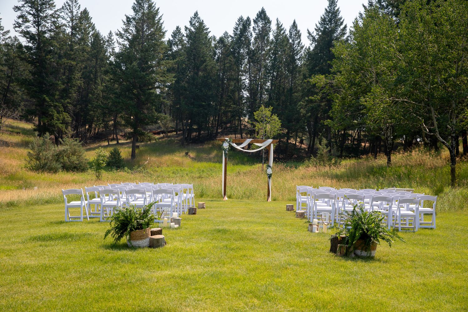 Outdoor wedding ceremony setup with white chairs, wooden arch, and lush green meadow surrounded by pine trees at Swansea Ranch in Invermere