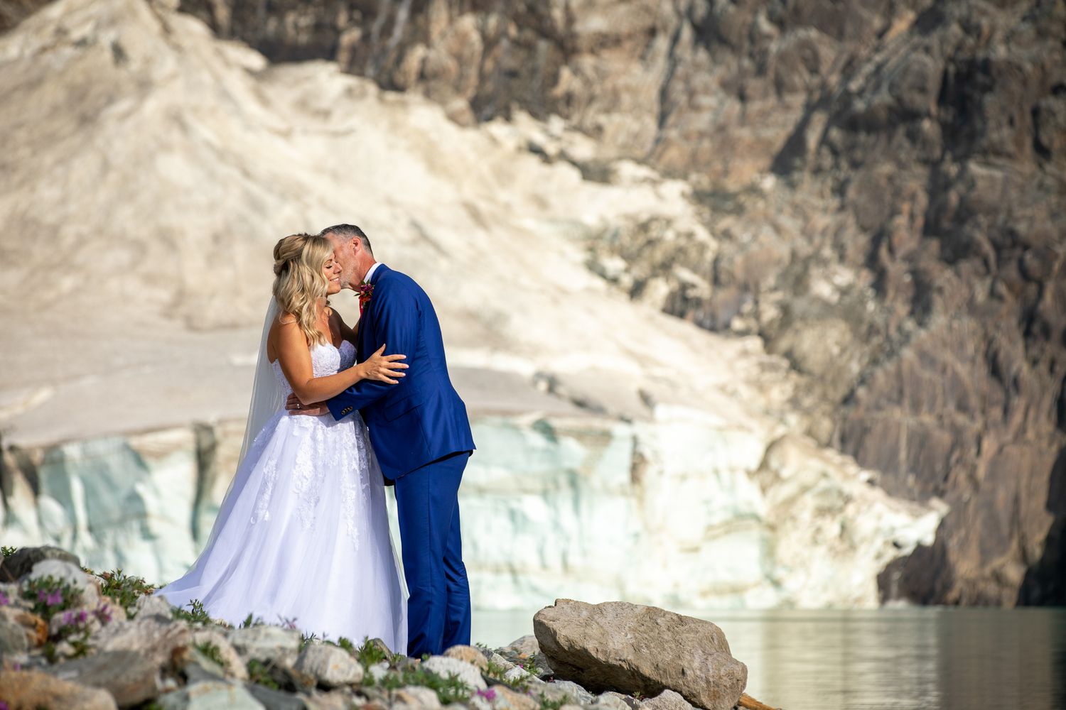 Bride and groom share a romantic kiss in wedding attire near a glacier and mountain backdrop. Copperpoint resort,  Invermere.
