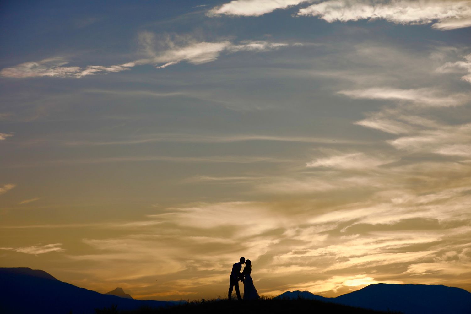 Two silhouetted wedding figures embrace against a dramatic sunset sky with mountains in the background. Swansea Ranch in Windermere, BC.