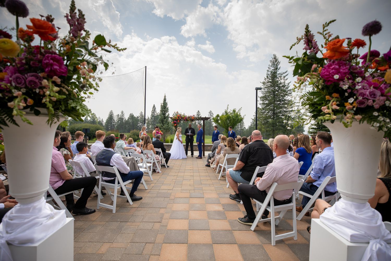 Outdoor wedding ceremony on a patio with guests seated in white chairs, colourfu floral arrangements, and couple at the altar. Invermere, BC