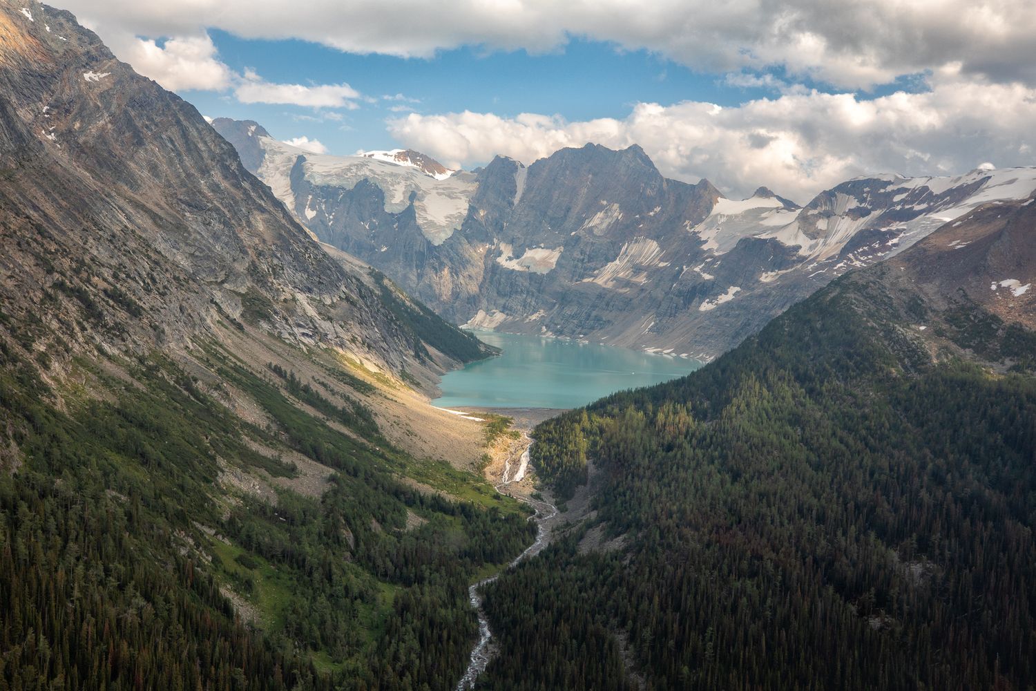 Aerial view of a turquoise mountain lake surrounded by pine forests and rocky peaks under a partly cloudy sky. Lake-of-Hanging-Glacier BC.