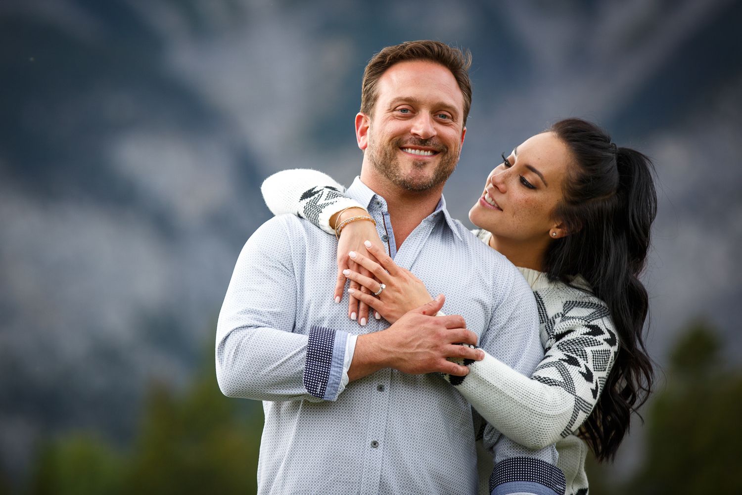 A smiling couple embracing outdoors against a cloudy sky background at Lake Minnewanka in Banff.