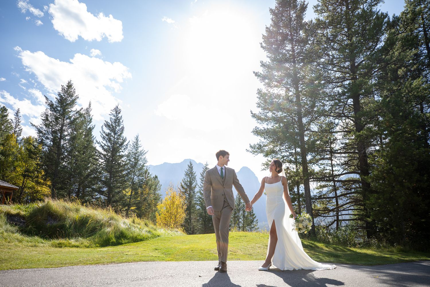 A couple in wedding attire stands on a scenic mountain overlook surrounded by evergreen trees and dramatic peaks. Silvertip resort, Canmore.