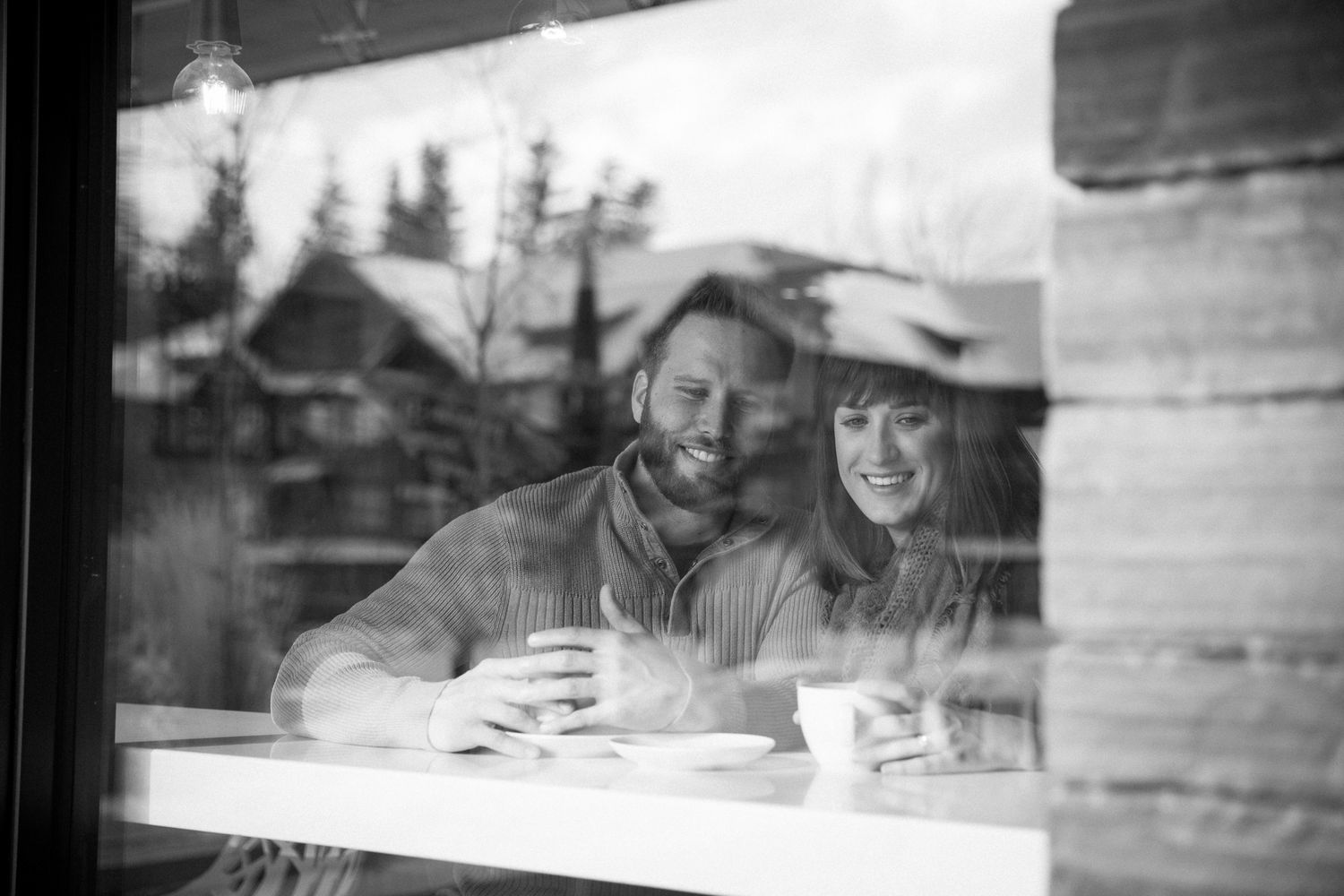 A black and white photograph shows two people smiling together at a table seen through a window reflection at Whitebark cafe in Banff, AB.