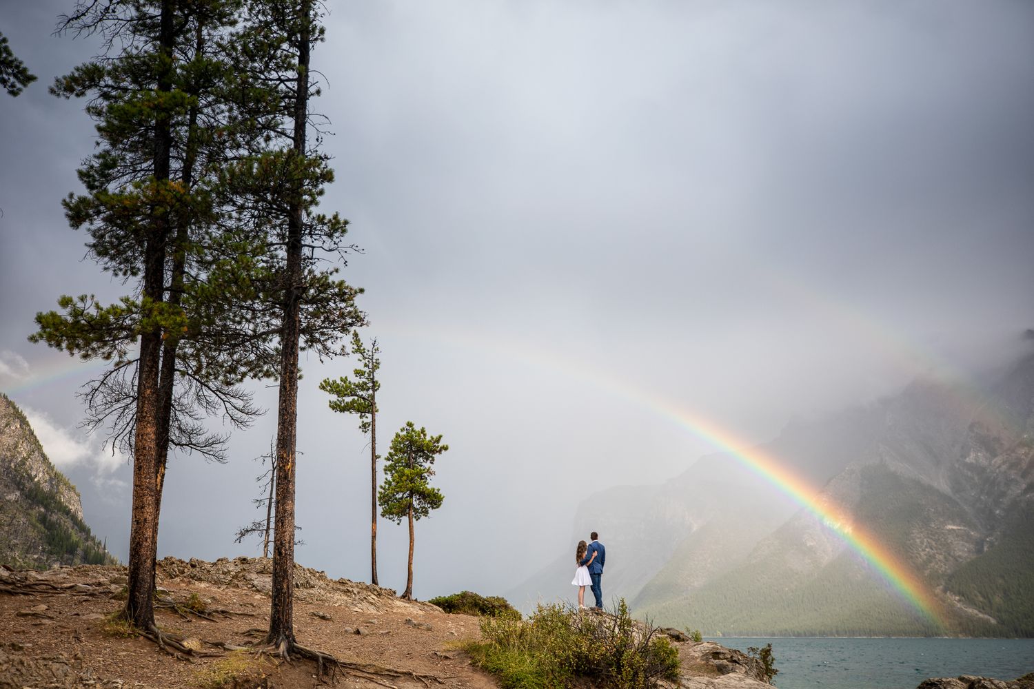 A rainbow appears over Minnewanka Lake in Banff a misty mountain landscape with tall pine trees silhouetted against dramatic storm clouds.