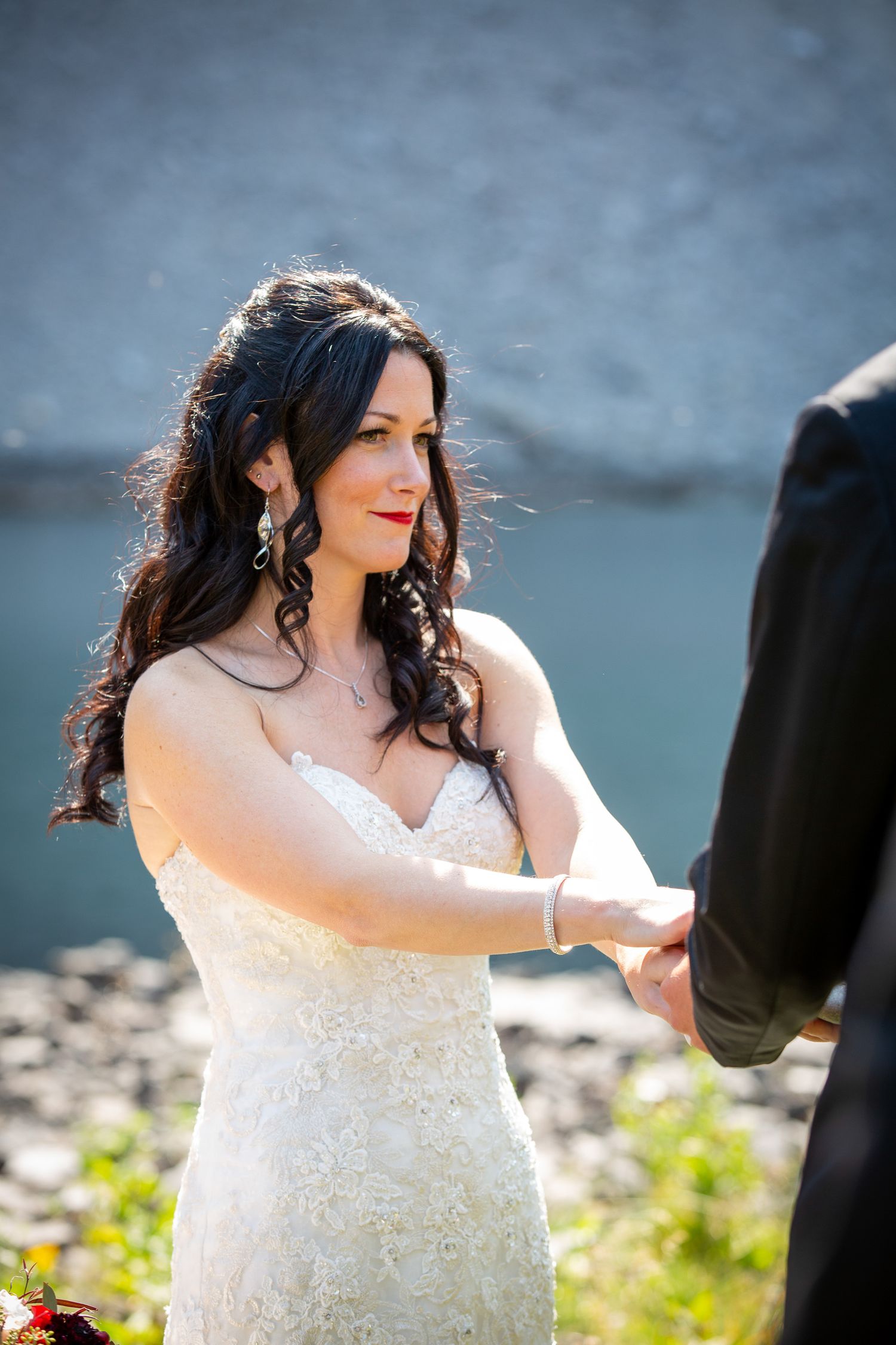 Bride in white lace gown holds groom's hands during outdoor wedding ceremony near a blue lake in Kananaskis, Alberta.