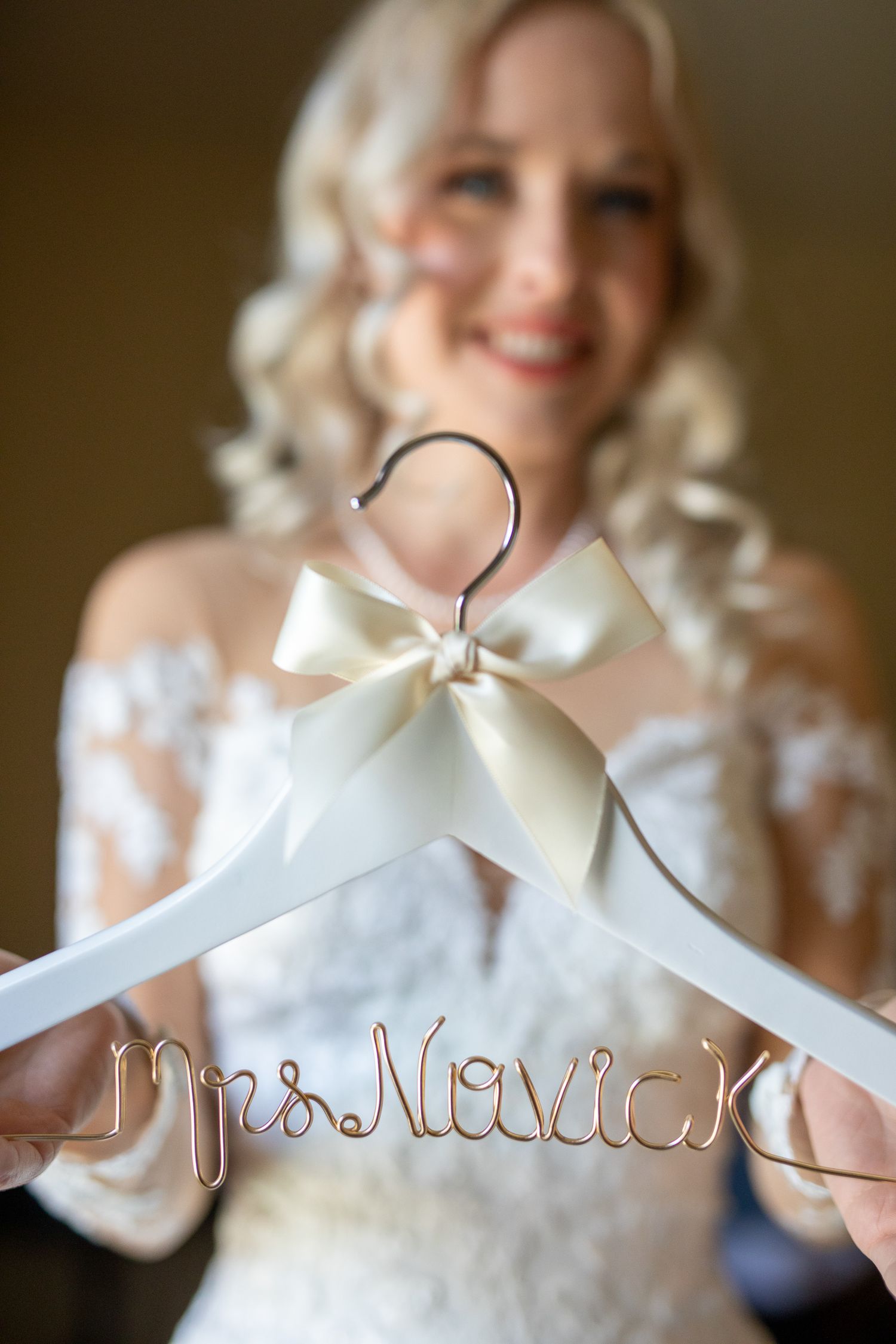 Smiling bride in lace dress holds custom wire name hanger reading Mrs Novick with ivory bow In Canmore