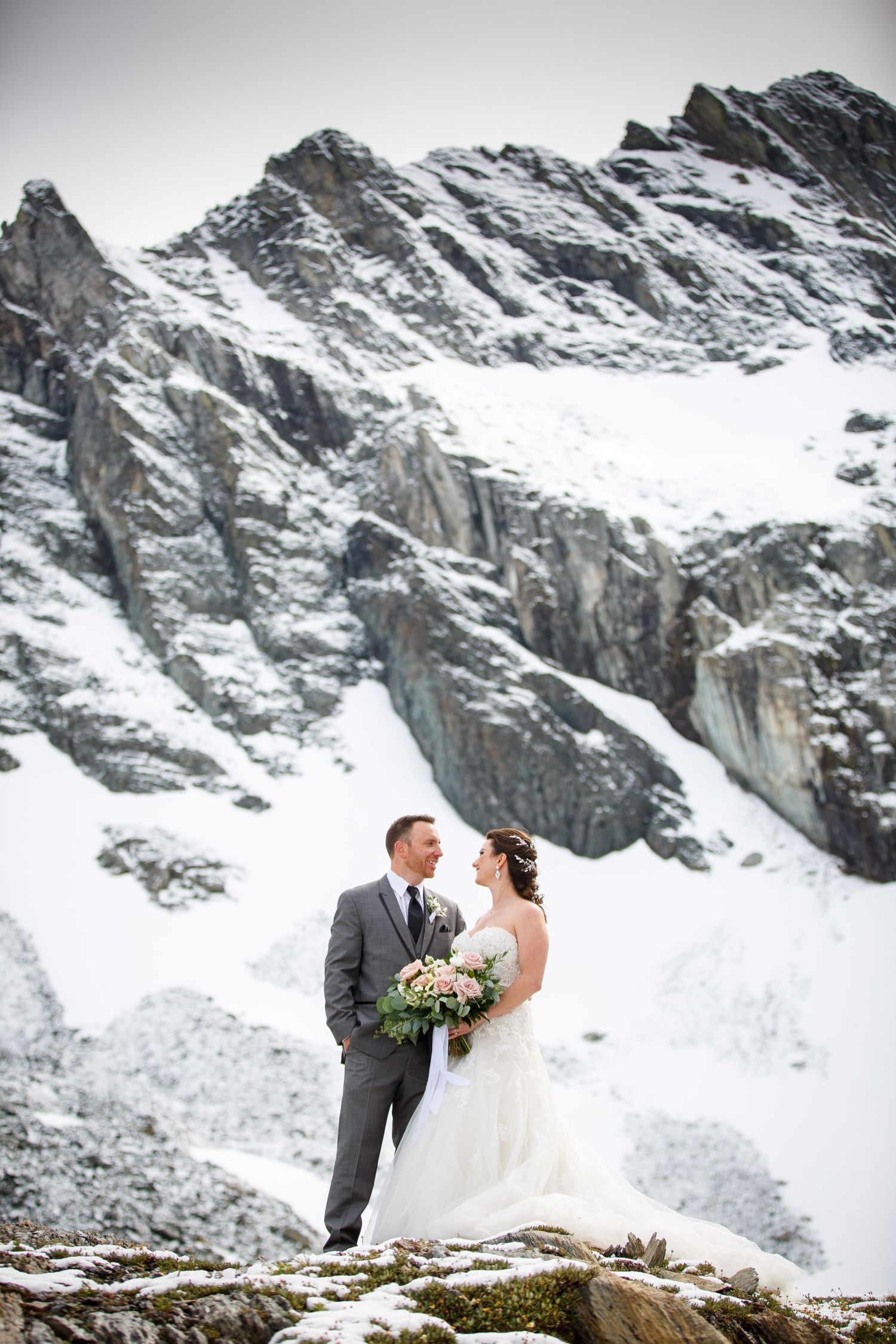 Bride and groom stand together on snowy mountain rocks, bride holding bouquet, groom in grey suit, stunning wedding backdrop Kimberley, BC