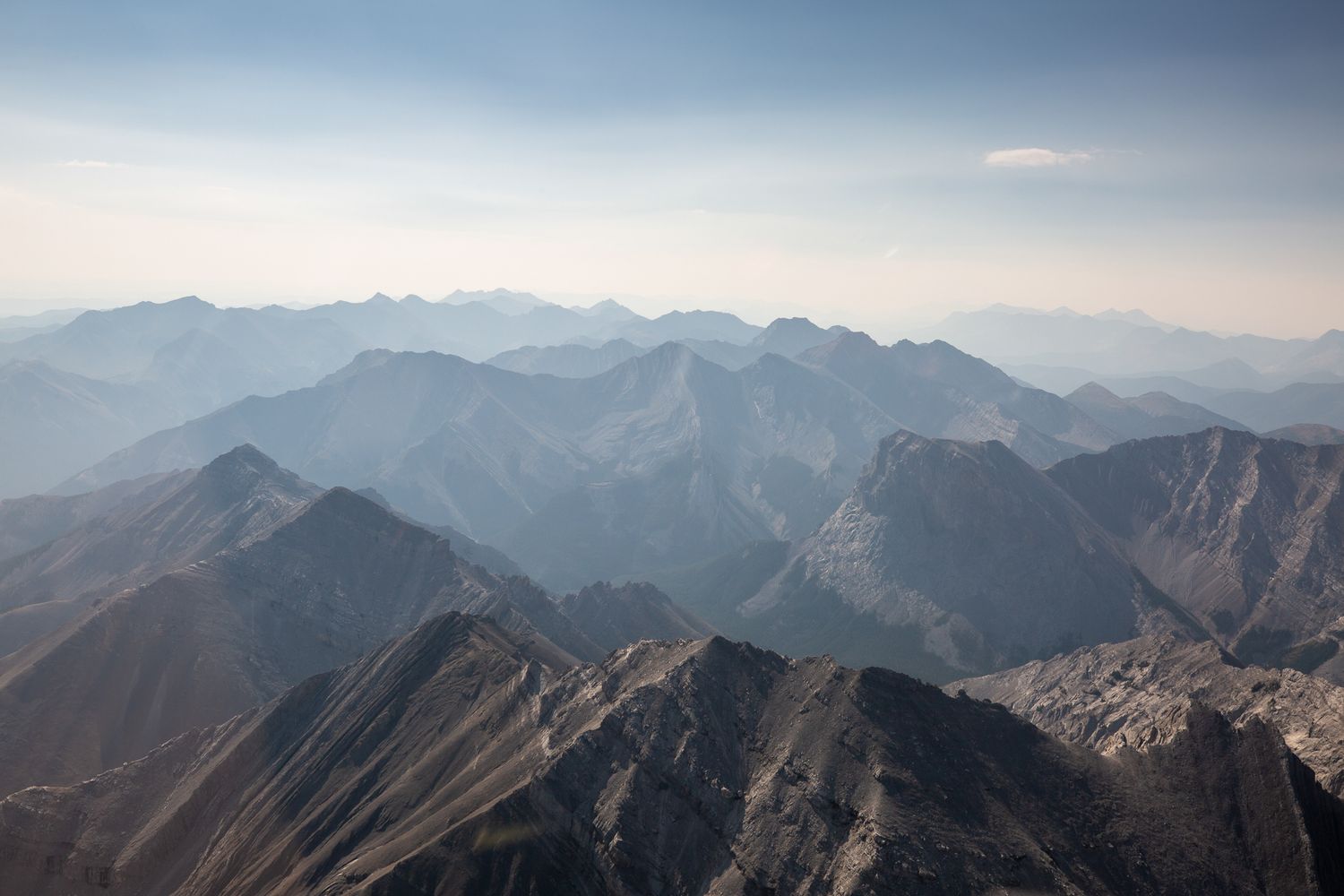 Aerial view of layered mountain ranges fading into a hazy blue horizon under a pale sky over in Kananaskis, Alberta.