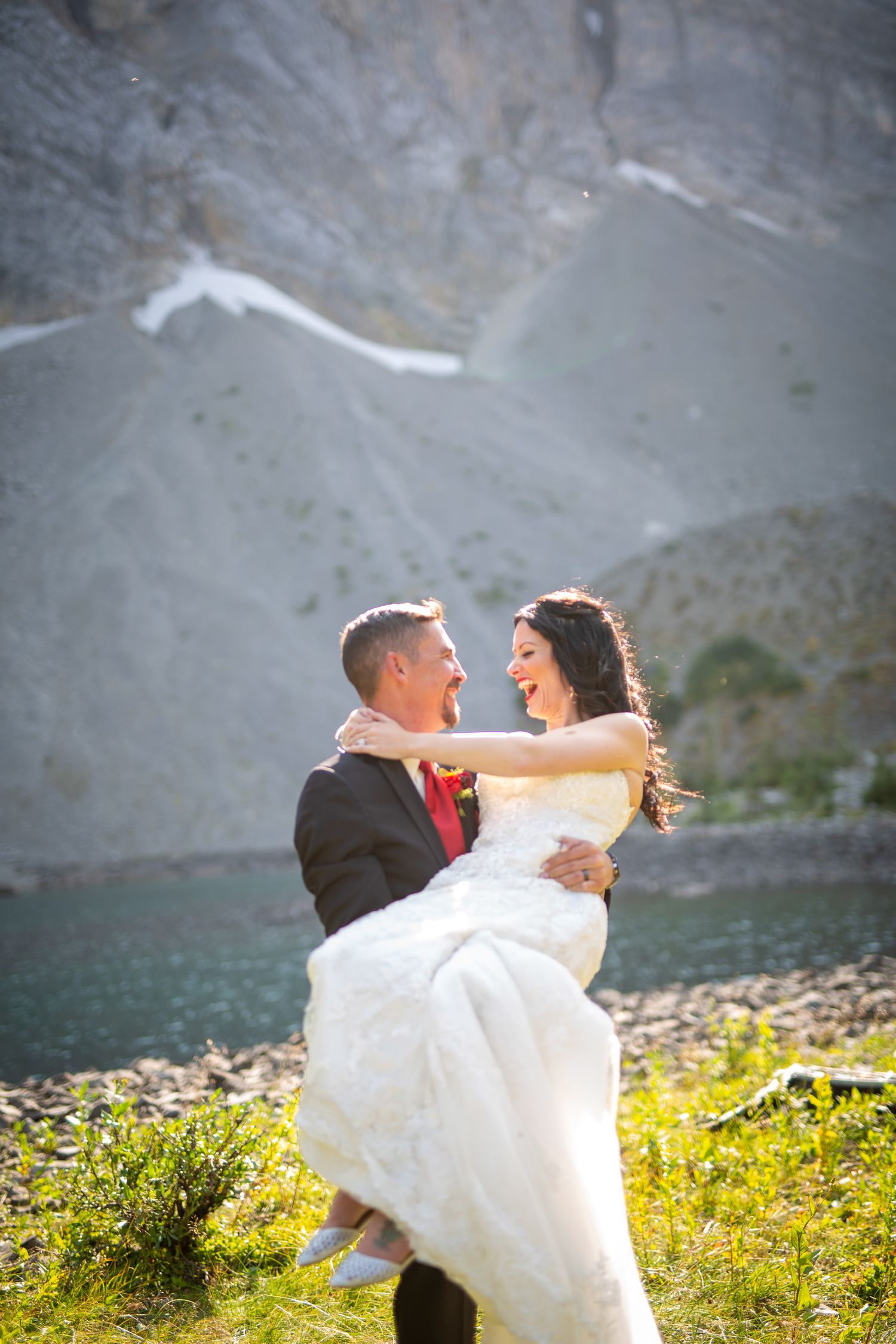 Groom carries laughing bride in white dress near mountain lake on sunny wedding day in Kananaskis, Alberta.