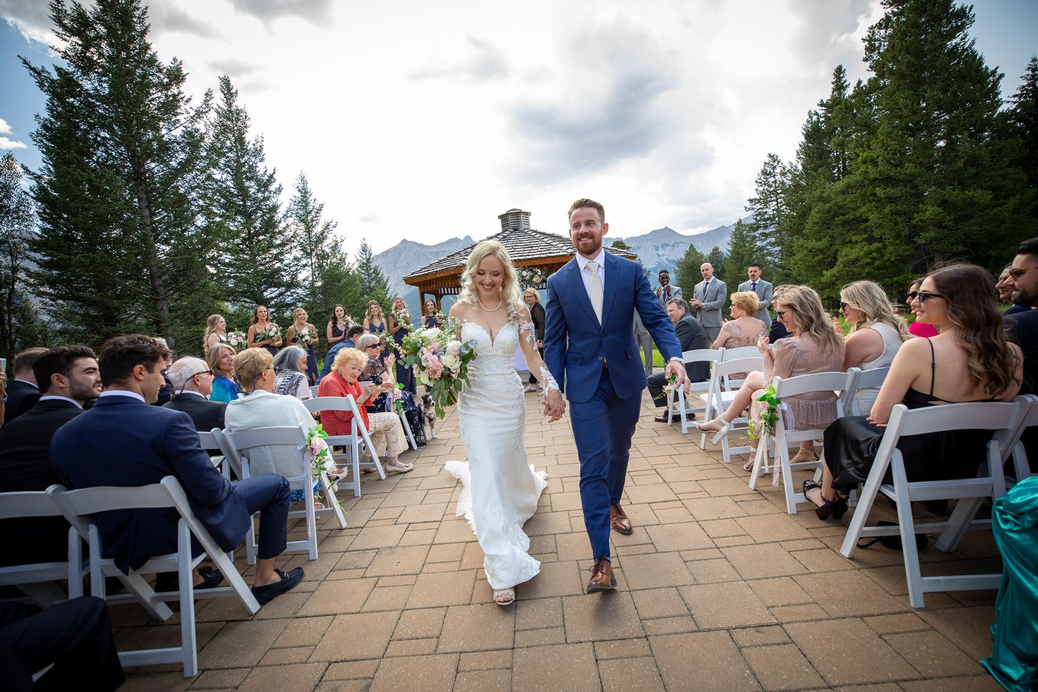 Bride and groom walk hand-in-hand down the aisle at an outdoor mountain wedding ceremony with seated guests at Silvertip gazebo in Canmore.