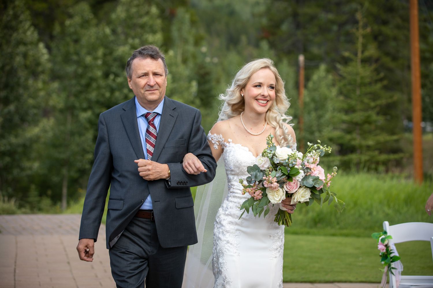 Bride in white gown holding bouquet walks arm-in-arm with escort at outdoor wedding ceremony In Canmore Silvertip gazebo.