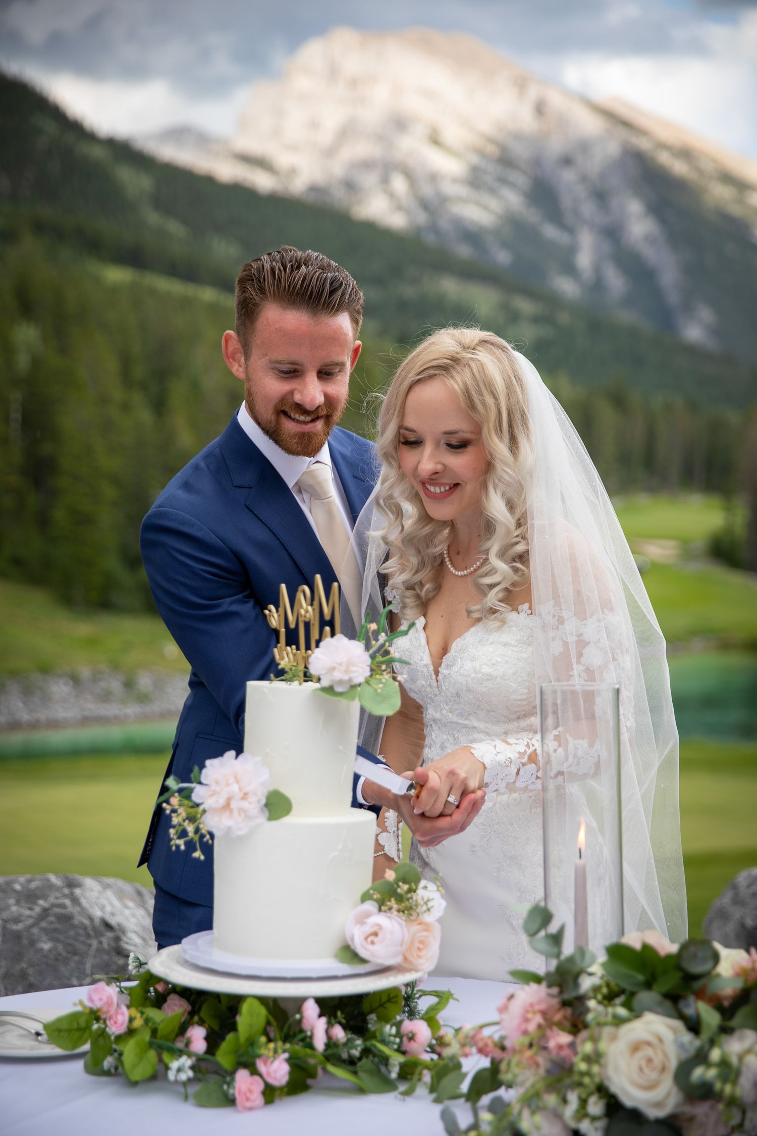 Bride and groom cutting white two-tier wedding cake outdoors with mountain and green forest backdrop at Silvertipin Canmore.