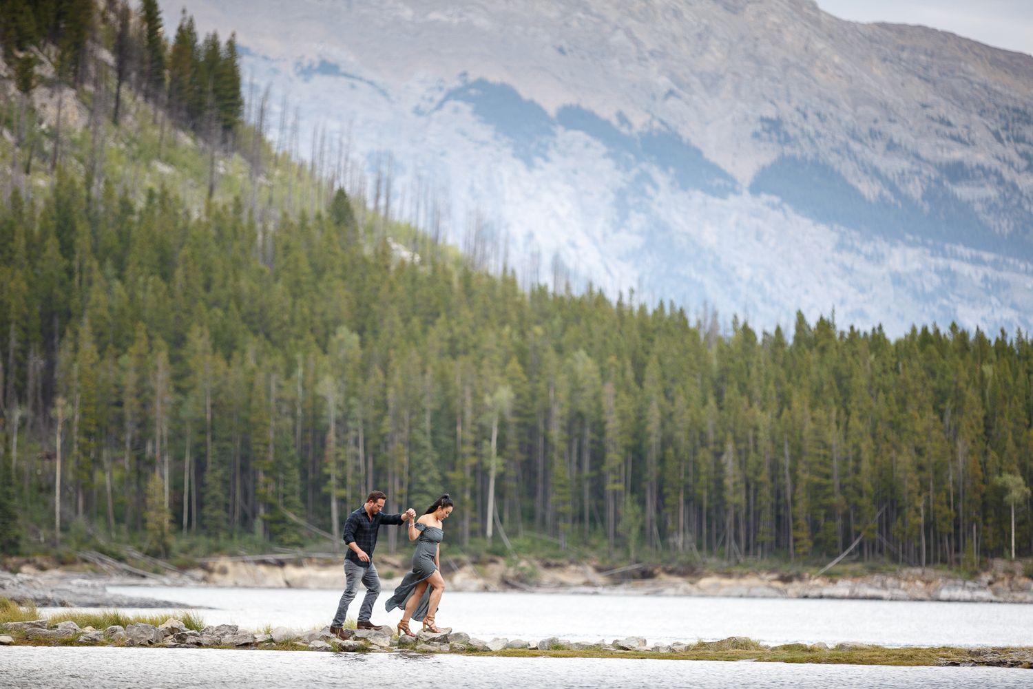 Two people walking on a rocky riverbank surrounded by a forested mountain landscape at Lake Minnewanka in Banff.