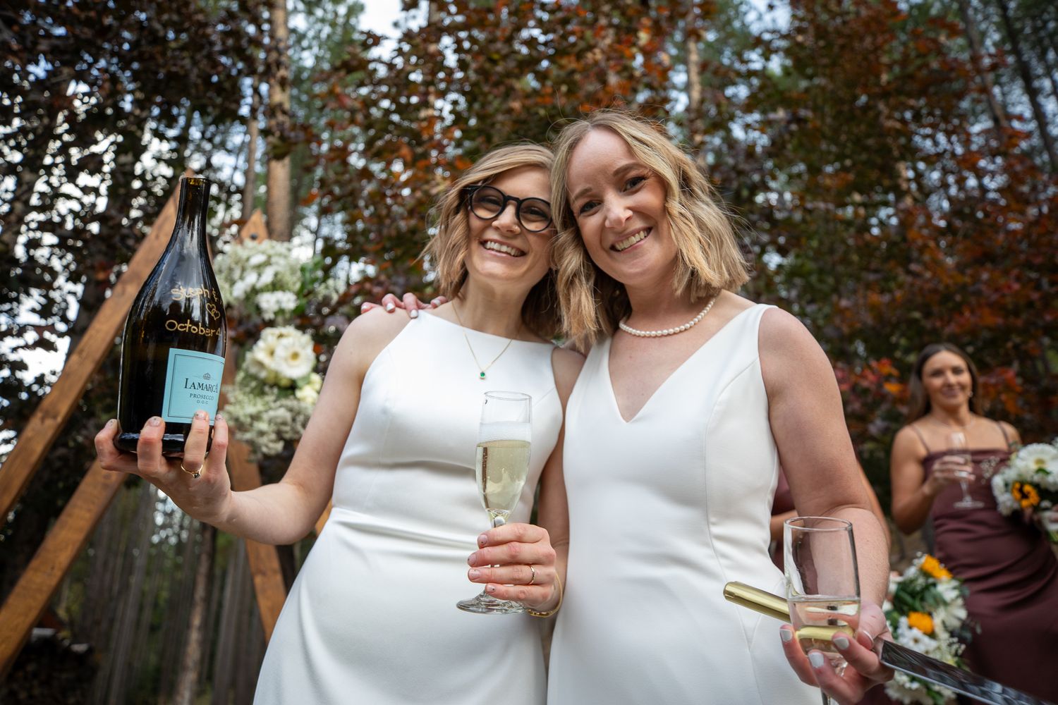 A LGBTQ couple in white dresses share a joyful moment during an outdoor celebration with autumn trees in the background. Earl Grey Lodge BC