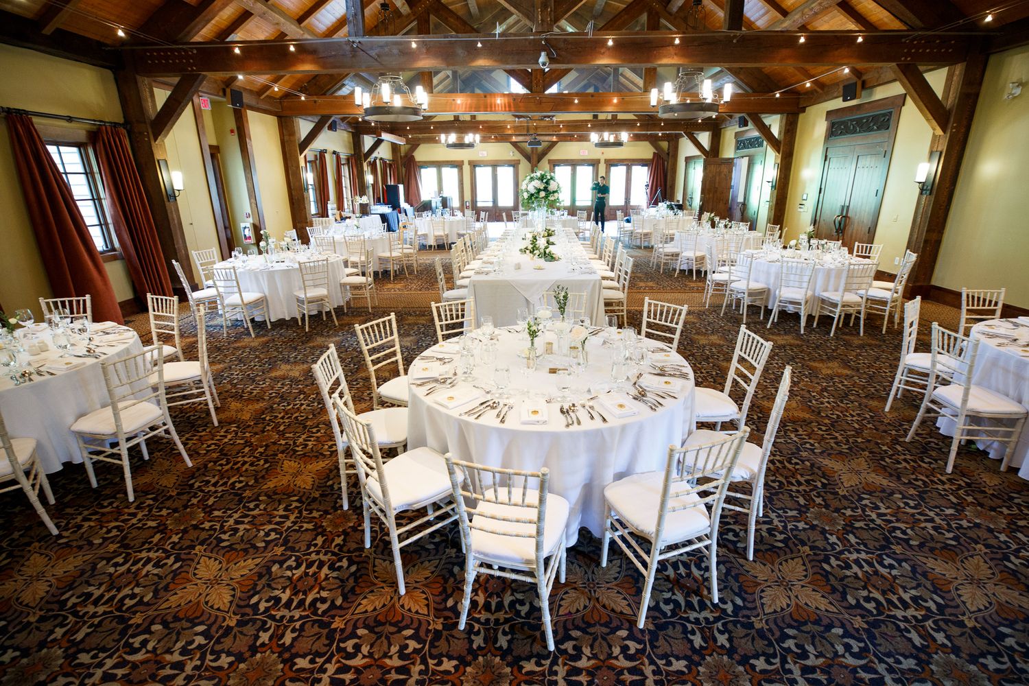 Elegant wedding reception venue with exposed wooden beams and round tables set with white linens and silver chiavari chairs. Silvertip.