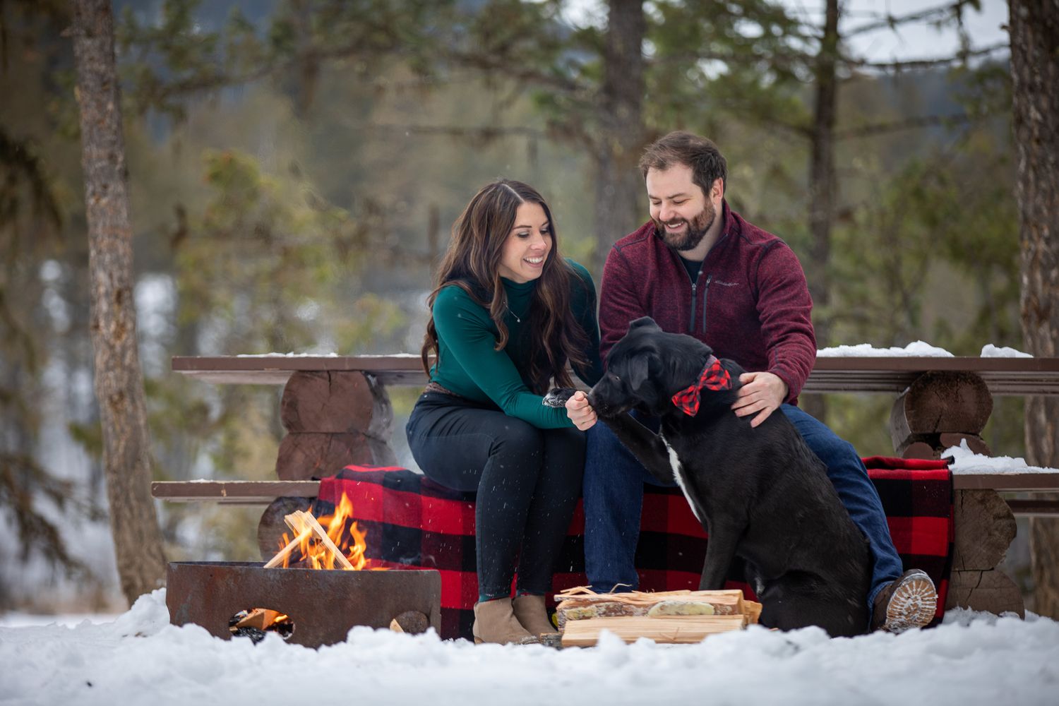 Couple sitting by a campfire in a snowy winter forest enjoying hot drinks and conversation at Grey Wolf Panorama, BC