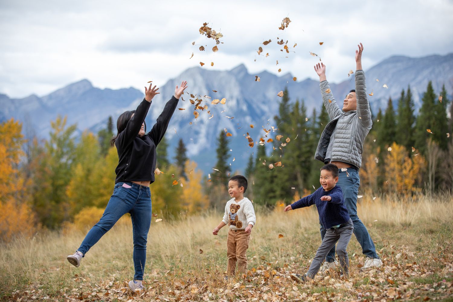 A family plays in fallen autumn leaves with mountains and evergreen trees in the background on a sunny day at Quarry lake park in Canmore.