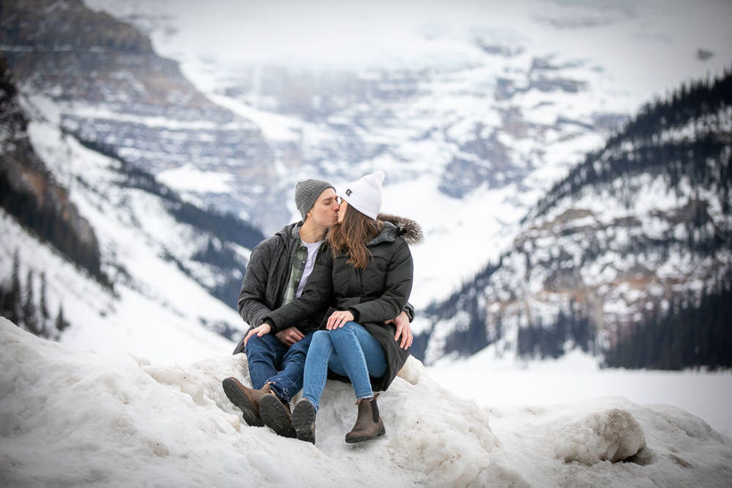 A couple sits on a snowy mountain ledge overlooking a dramatic winter Lake Louise landscape in the Canadian Rockies.