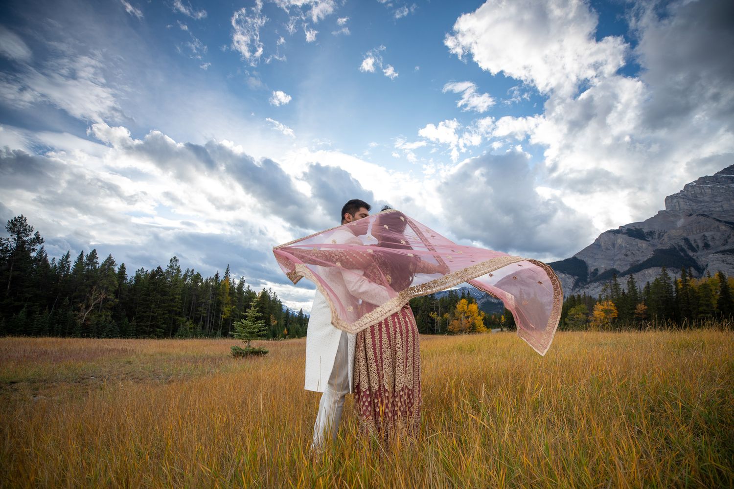 A flowing pink dress billows in the wind against a dramatic mountain landscape with cloudy skies in the Bumpy meadow in Banff, Alberta.
