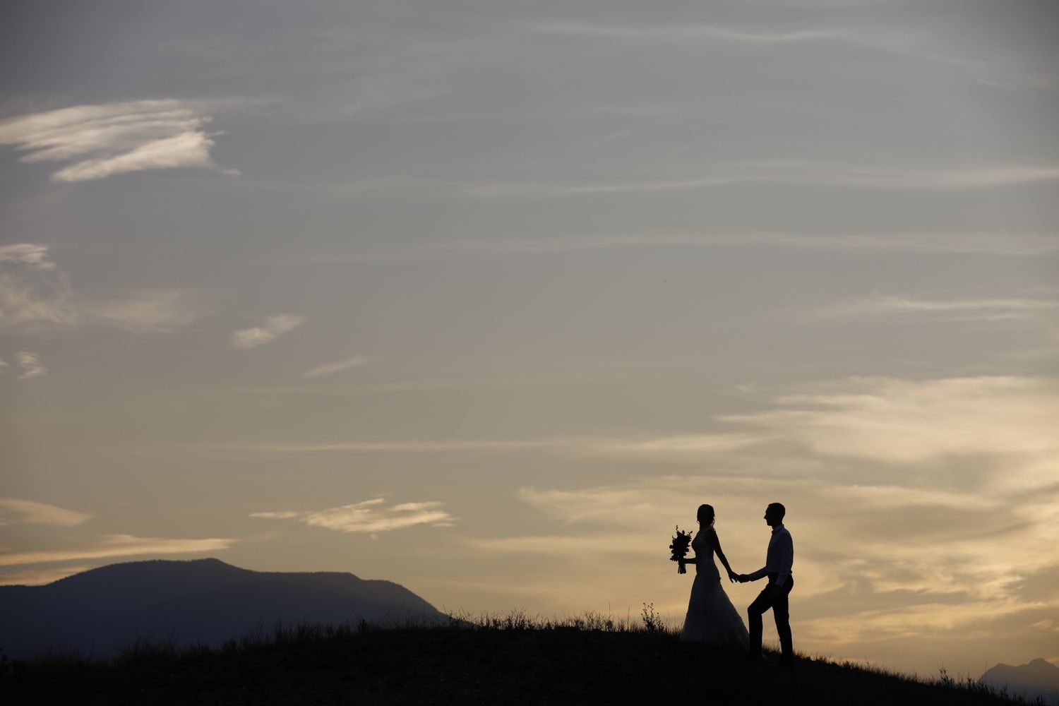 Silhouetted couple stands on hilltop against dramatic golden hour sky with mountains in the background at Swansea Ranch in Windermere, BC